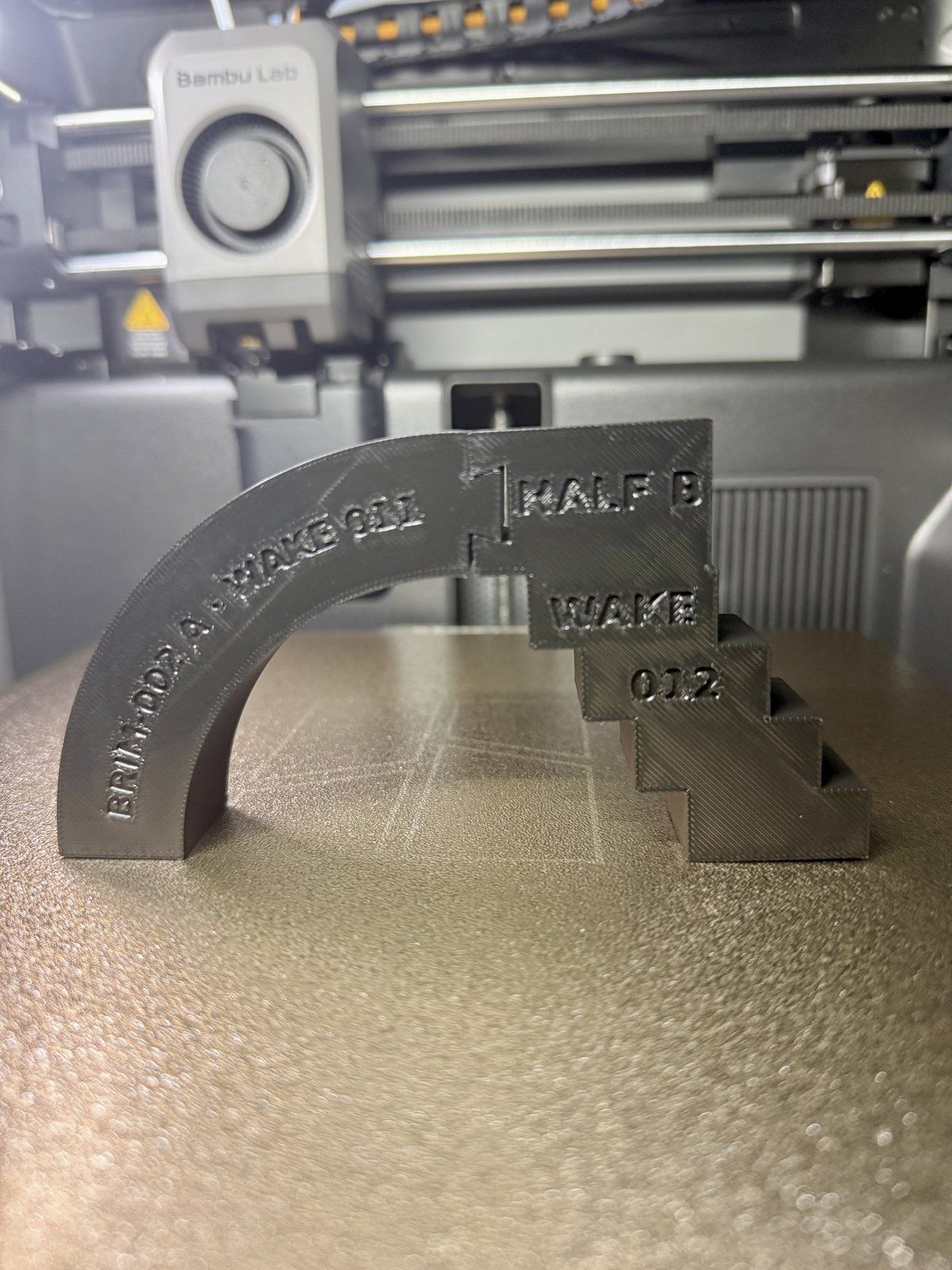 Photograph of BRIM-002 assembled on the printer bed: half A's smooth quarter-arch curve, debossed 'BRIM-002 A · WAKE 011', rises from the left and meets half B's corbelled stair, debossed 'HALF B / WAKE / 012', at a visible dovetail joint at the crown. The arch stands freely with nothing supporting it.