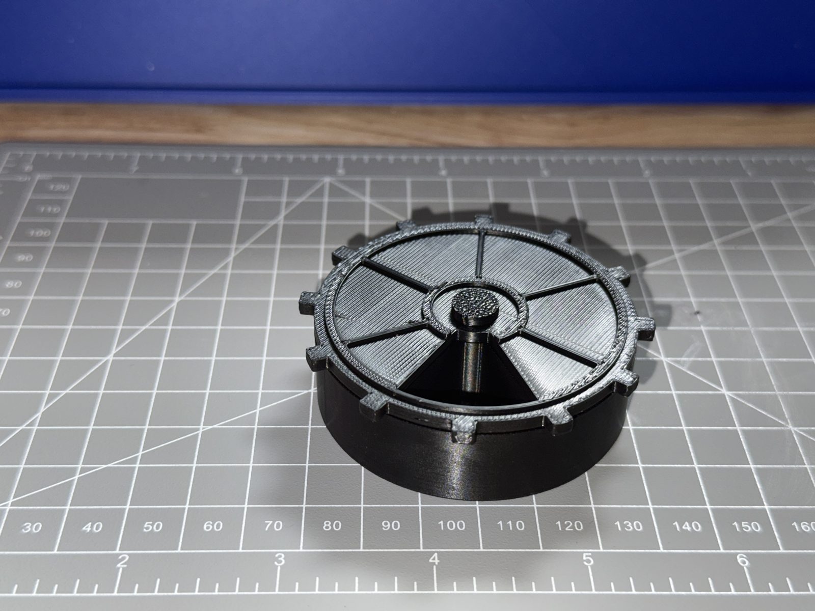 The printed set assembled on a cutting mat: black PETG cup, grey vault-door lid with twelve gear teeth seated on it, pin standing proud in the centre.